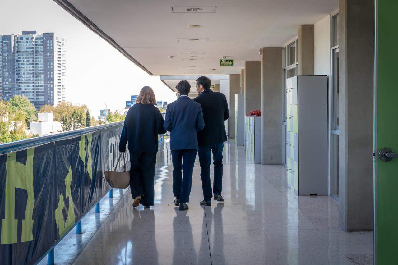 Tres personas caminan por un corredor moderno de la escuela hacia una reunión, con vista a la ciudad de fondo.